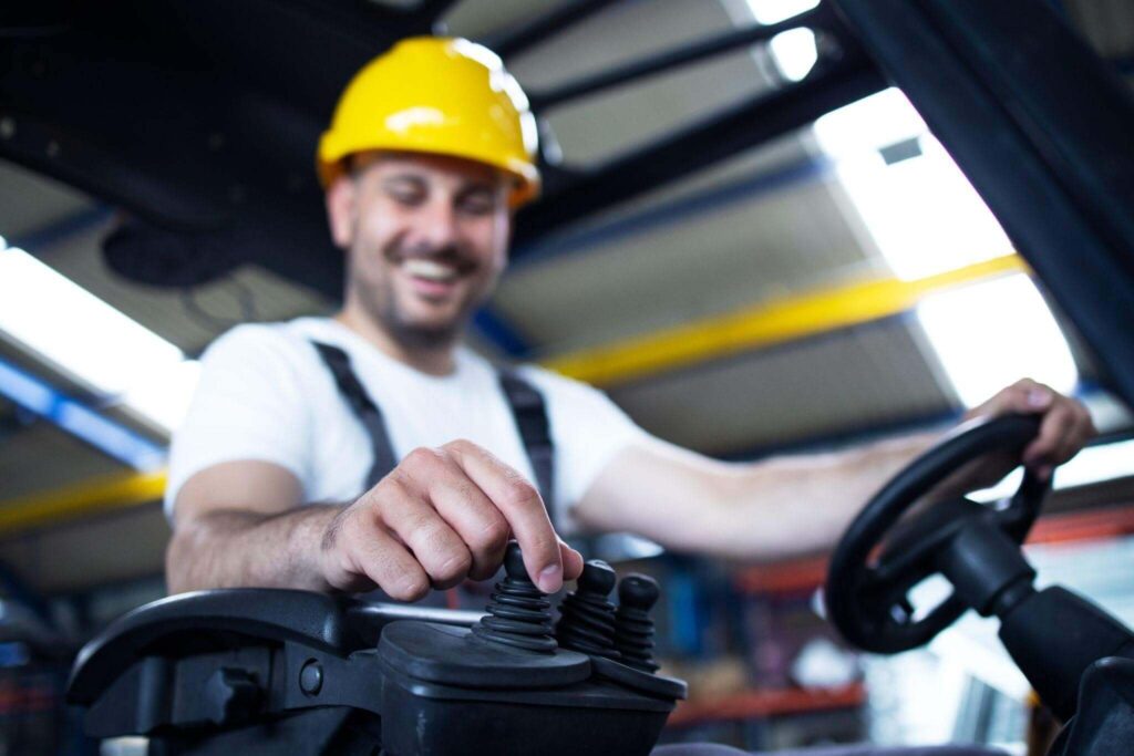 Foto em plano médio e ângulo ligeiramente inferior focando na mão de um homem que trabalha como operador de empilhadeira. Ele está manuseando as alavancas de controle do veículo, com o volante visível ao lado. O homem está sorrindo em segundo plano, ligeiramente fora de foco, vestindo uma camiseta branca, macacão de trabalho escuro e um capacete de proteção amarelo. O cenário é o interior de um armazém.