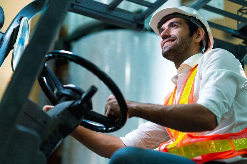 Vista lateral em ângulo inferior de um homem sorridente trabalhando como operador de empilhadeira dentro de um centro de distribuição. Ele segura o volante do maquinário enquanto olha para cima e para a frente. O profissional veste uma camisa branca de botões, um colete de segurança reflexivo nas cores laranja e amarelo, e utiliza um capacete de proteção branco. A estrutura metálica da cabine e o ambiente do estoque compõem o fundo.